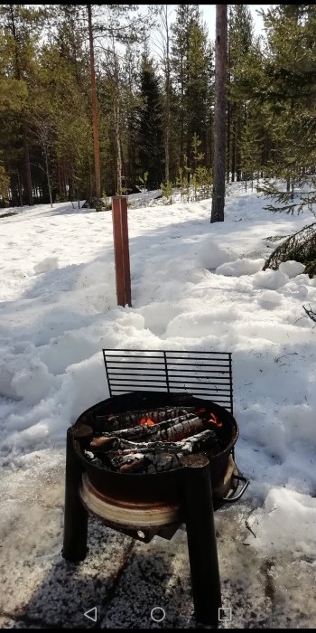 En grill fylld med glödande kol står i snötäckt landskap med skog i bakgrunden.