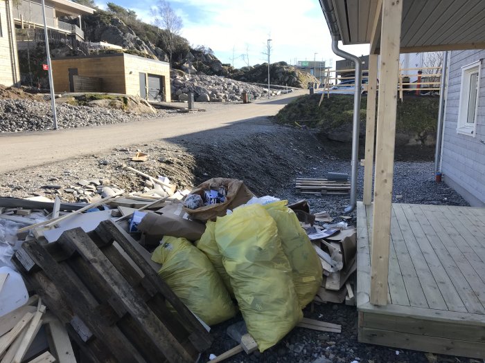 Grusig slänt framför hus med byggavfall nära träveranda, i bakgrunden syns en gata och berg.