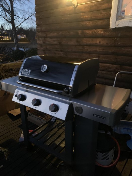 En svart och silverfärgad gasolgrill av märket Weber Genesis II placerad på en trätrall utomhus vid skymningen.