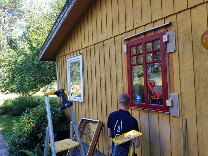 Person som byter fönster på en gul träfasad, rött fönster och verktyg på stege.