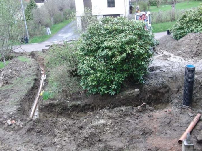 Grävmaskin vid pågående dräneringsarbete i trädgård med synliga jordhögar och vegetation.