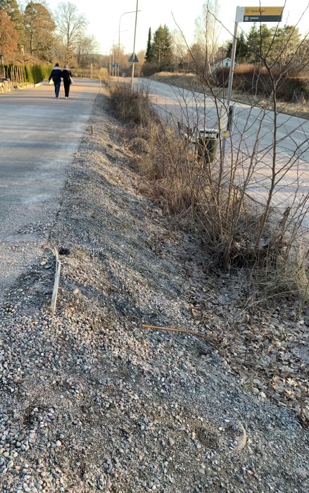 Slänt vid tomtgräns med grus och lite vegetation, intill en asfalterad väg där två personer går.