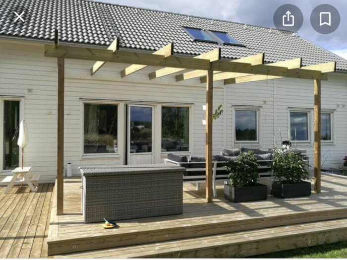 Nybyggd trädäck med pergola i trä vid en vit villa, möblerat med loungesoffor och planteringslådor.