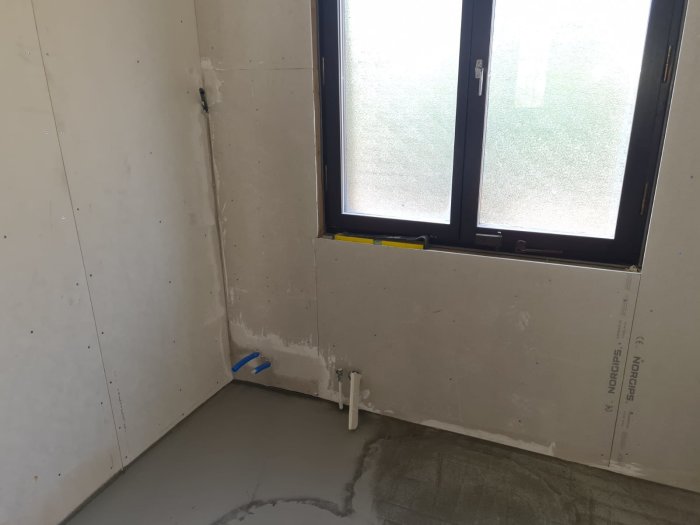 Renoveringsarbete i badrum med väggplattor, rörinstallationer vid fönster och vattenanslutningar på golvet.