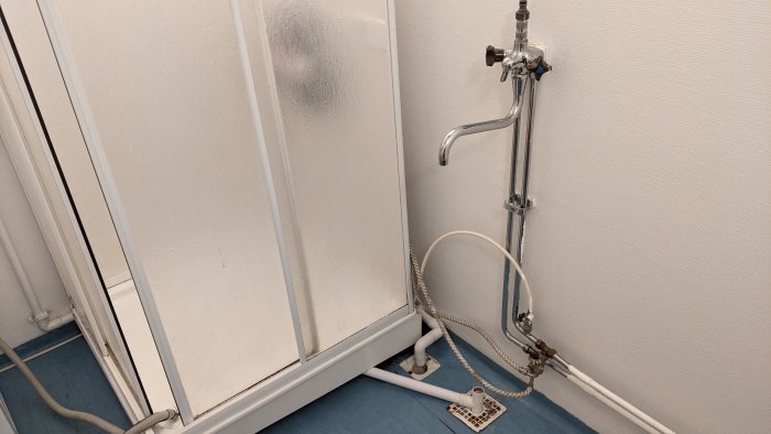 Hörn av ett äldre badrum med duschkabin, synliga rör och vattenkopplingar på väggen.