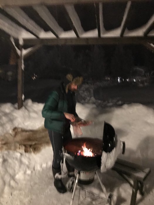 Person som grillar utomhus i snö med en kolgrill på kvällen.