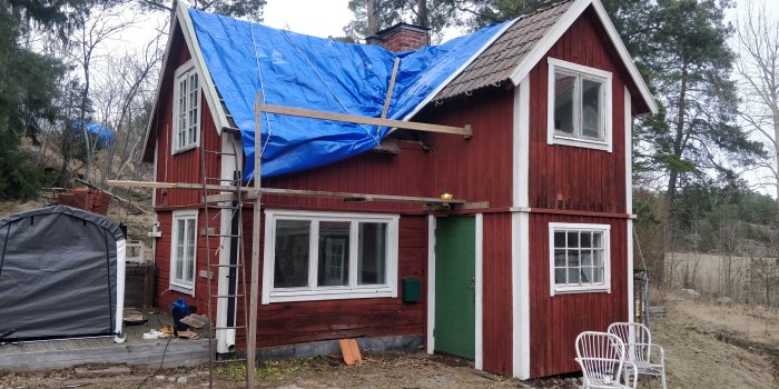 Rött hus med blå presenning på taket under renovering, ställningar och utspridda byggmaterial runtomkring.