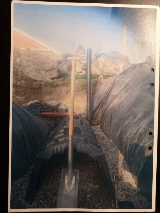 Nedgrävd markbädd med synligt svart dräneringsrör, grus och spade, intill stora stenar och grönt staket.