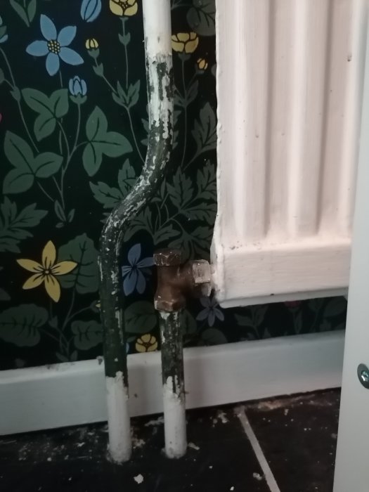 Vattenrör och gammal termostat vid en vägg med blommigt tapetmönster, bredvid ett vitt radiatorselement.