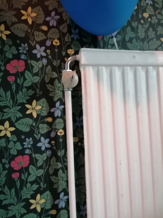 Vänster sida av radiator med inkommande vattenrör och termostat mot blommig tapetbakgrund.