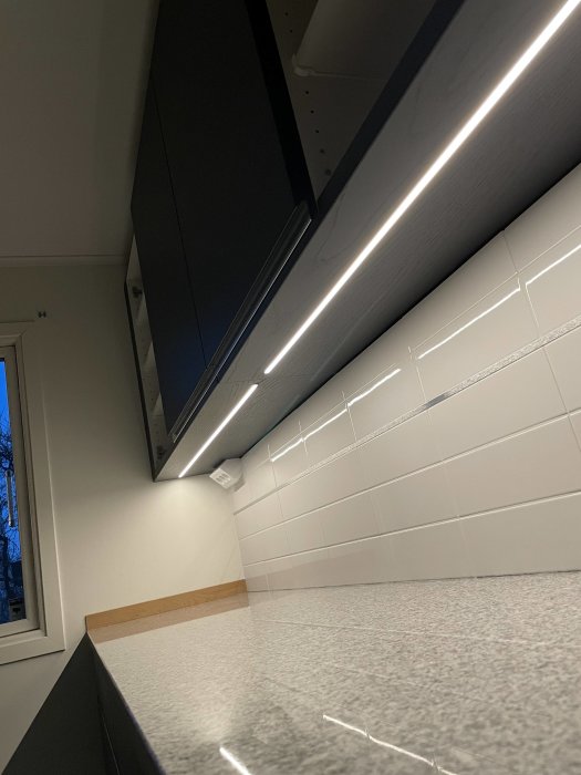 Kök med grå bänkskiva, vita kakelväggar och LED-lister under överskåpen.