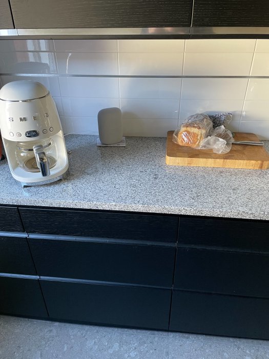 Nytt kök med svarta skåpluckor, vit kakelvägg, bänkskiva i sten och köksassistent på bänken.