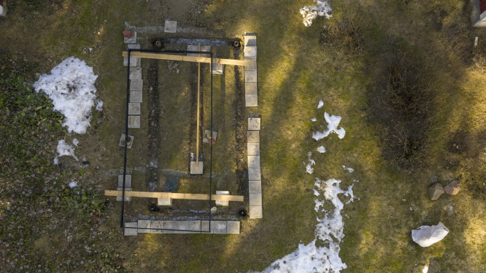 Flygbild av förberedd grund med aluminiumram för gjutning i en trädgård, omgiven av gräs och smältande snö.