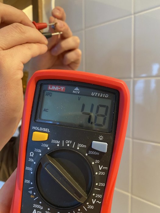 Hand håller en multimeter som visar 4.8 volt mot kaklad väggbakgrund.