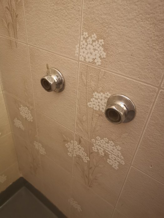 Två öppna röranslutningar på en vägg med blommigt kakel, möjligtvis för ett tidigare badkar.