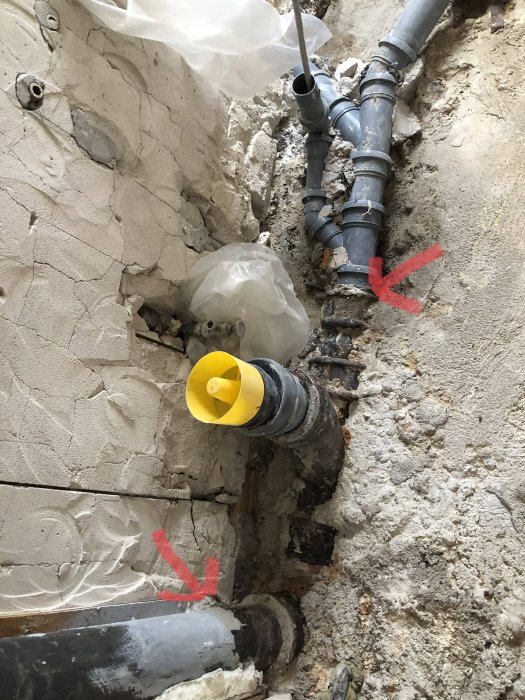 Felaktig anslutning mellan gammalt gjutjärnsrör och nytt plaströr i en vägg under renovering, markerat med röda pilar.