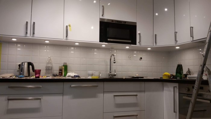 Renoverat ljust kök med vita skåp, spotlights, kakel, rörigt arbetsbänk och stege till vänster.