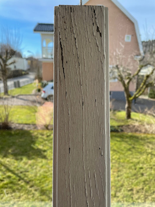 Närbild på en fönsterbåge med flagande färg och sprickor i träet mot en suddig bakgrund av en trädgård.