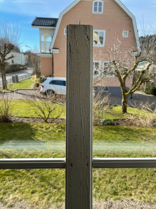 Ett träfönster sett inifrån, med färg som släpper i nedre delen, i ett hus med en trädgård och annat hus i bakgrunden.