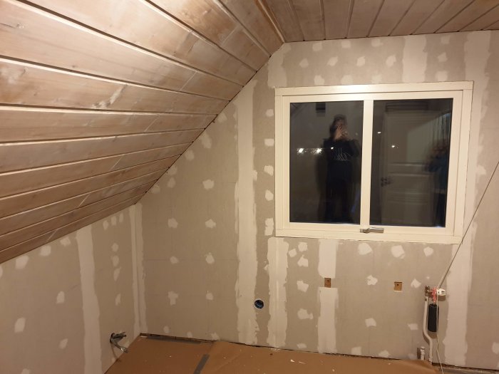 Renovering av sovrum med halvfärdiga gipsskivor och träpanel på en snedtaksvägg, delvis spacklad vägg och fönster.