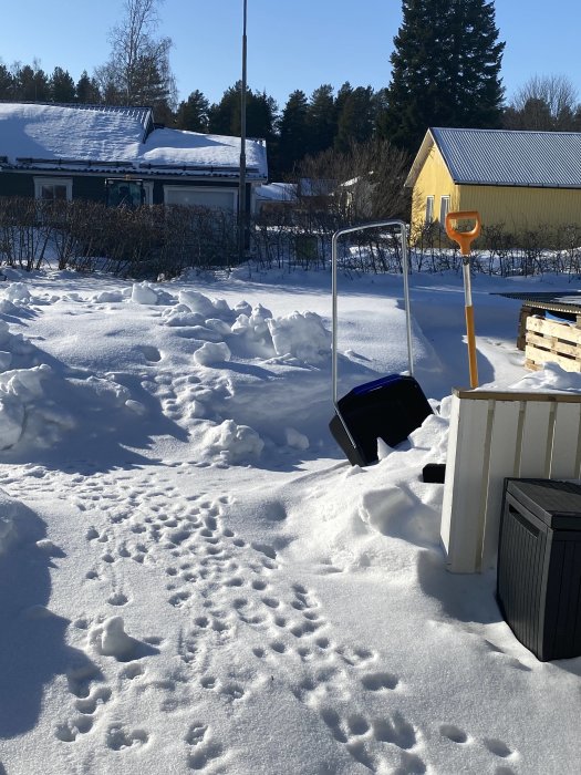 Skottkärra i snötäckt trädgård med spår i snön, omgiven av snödrivor och hus i bakgrunden.