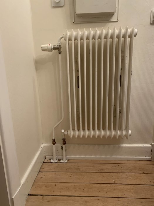 Vit radiator på en vägg ovanför ett trägolv i en hörna av ett rum, med rörledningar kopplade till golvet.