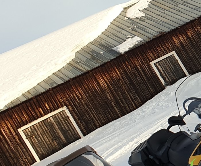 Snötäckt ladutak med isformationer på kanterna, brun trävägg och snöskoter delvis synlig.