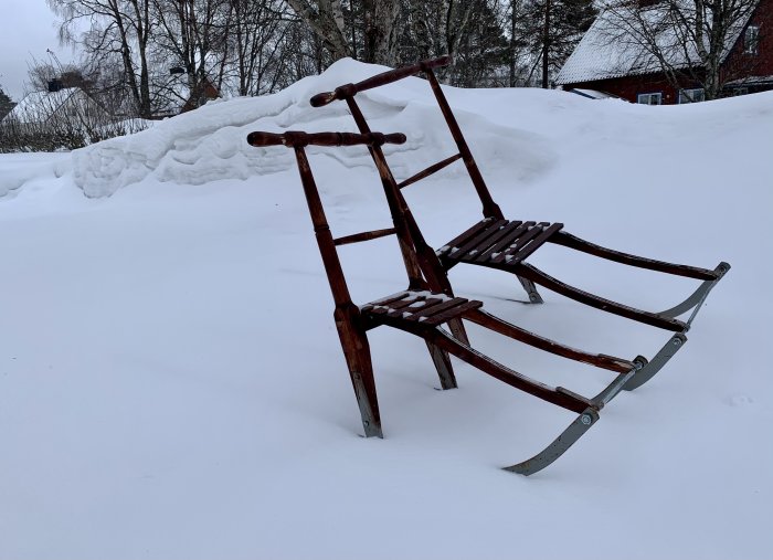 Träsläde med metallmedar i snö med snötäckta träd och rött hus i bakgrunden.