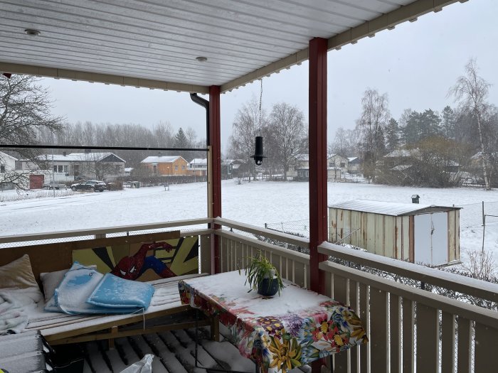 Öppen veranda med tak, räcken, och en soffa, med utsikt över en snötäckt trädgård, utan fönster eller värmekälla.