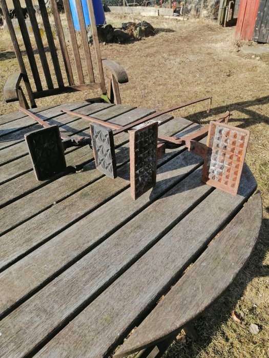 Rostiga smidesverktyg med mönstrade plattor på ett träbord utomhus.