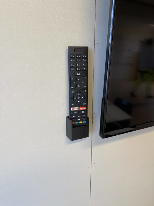 3D-utskrivet svart fäste för fjärrkontroll monterat på vägg vid en TV-skärm.