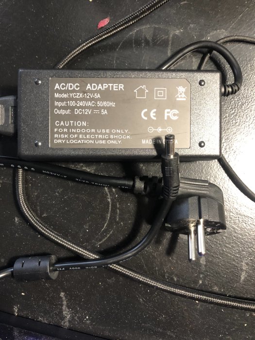 AC/DC-adapter och kablar mot en svart bakgrund, varningstexter och säkerhetsikoner synliga.