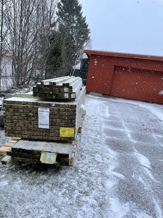 Byggmaterial staplat på pallar på en snötäckt uppfart med röd byggnad i bakgrunden.