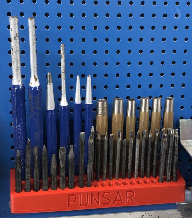Samling av olika storlekar på stansar och punsar i en orange hållare med texten "PUNSAR" framför en blå verktygstavla.