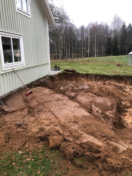 Uppgrävning för tillbyggnad intill tvåvåningshus på landet, med synligt berg och jordhög.