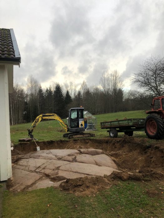 Grävmaskin bredvid en uppgrävd markyta med synligt berg i förberedelse för husbygge på landet.