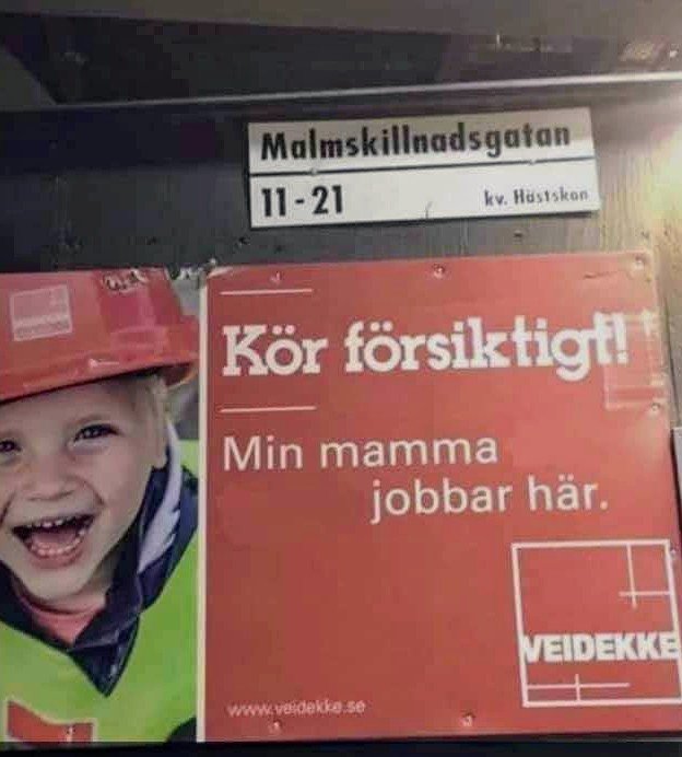 Skylt med texten "Kör försiktigt! Min mamma jobbar här." bredvid leende barn i bygghjälm.