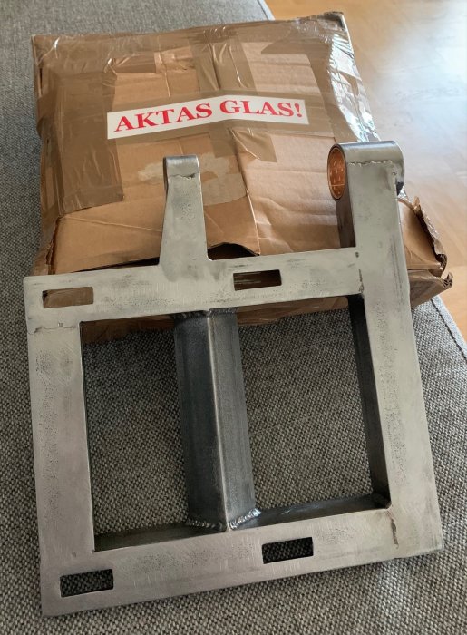 En metallram med en brun kartong märkt "AKTAS GLAS!" med tejp på en soffa.