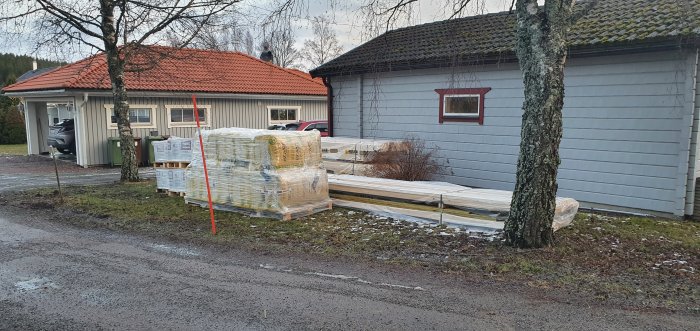 Byggmaterial packat och plastat på pallar framför en bostad med grått uthus och bil i carport.