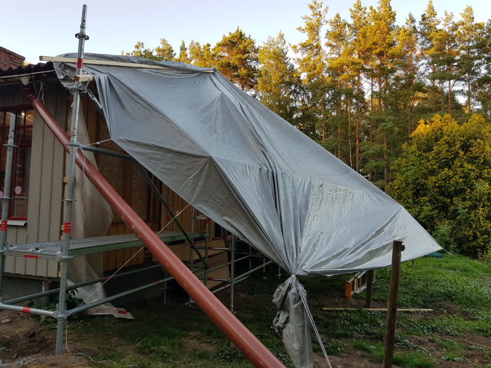 Tält uppställt över byggnadsställningar vid en husrenovering, omgivet av gröna träd.