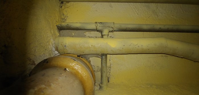 Äldre gulmålade avloppsrör mot en gul vägg i källaren, med fokus på kopplingen för badrumsrenovering.