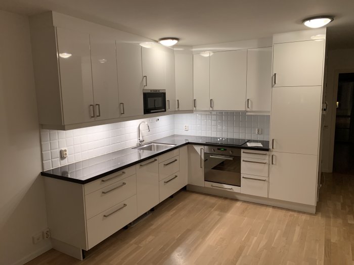 Ett nyrenoverat kök med vita skåp, rostfria handtag och svart bänkskiva, upplyst av spotlights.