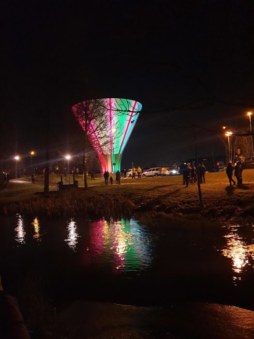 Vattenreservoar upplyst i neonfärger med reflektioner i vatten och mörk kvällshimmel.