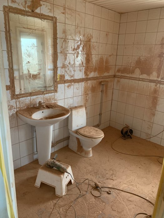 Renoveringsprojektets start i ett badrum med vita kakelväggar, smutsig tvättställ och toalett, byggdamm och verktyg på golvet.