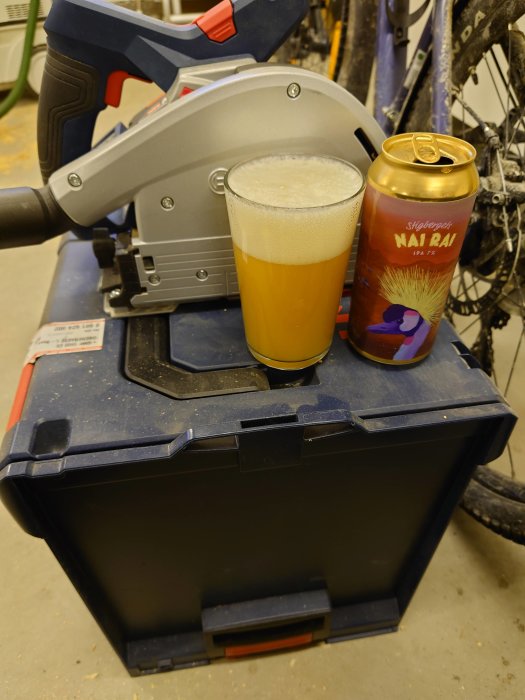 Ölglas med skum på verktygslåda bredvid en ölburk och en gersåg i en verkstad.