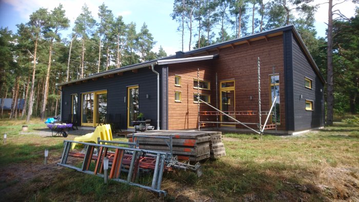 Trähus med fasad målad i svart och brun lasyr, med skog i bakgrunden och byggmaterial i förgrunden.