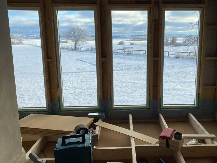 Utsikt från en pågående byggarbetsplats inne i hus med synliga träreglar och verktyg mot ett snöigt landskap.