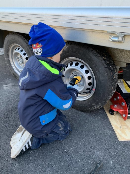 Barn i blå jacka använder en elektrisk skruvdragare på en bilhjulsmutter med röd domkraft nära.