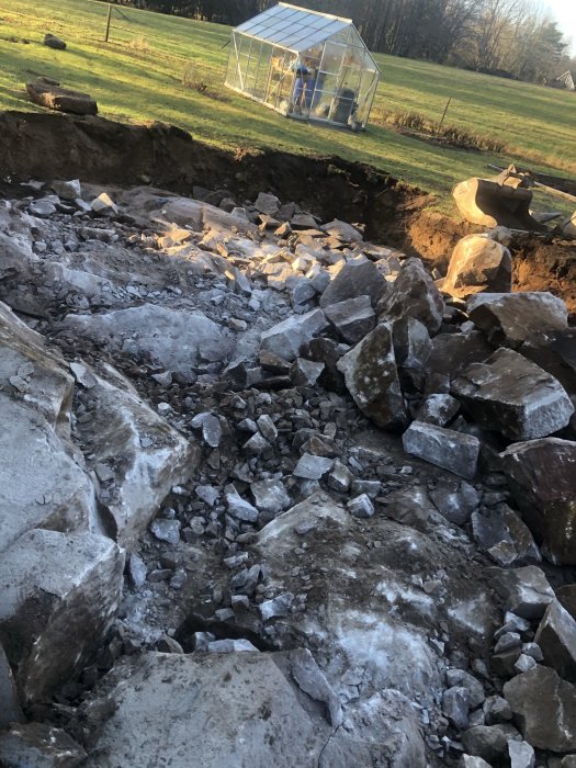 Stor hög med bruten sten på byggarbetsplats efter hydrospräckning, med växthus i bakgrunden.