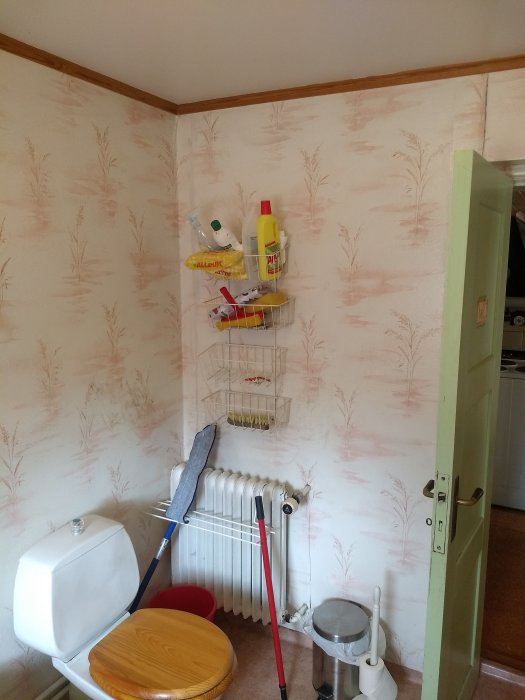Badrumsinteriör med toalett, radiator, tvättmedel på hylla, tvättkorg och städredskap i ett äldre hus.